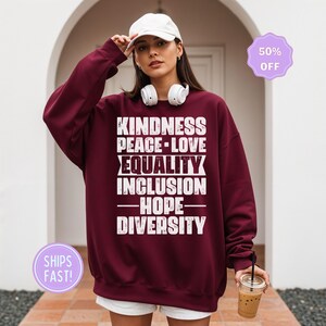 May include: A maroon sweatshirt with the words "KINDNESS PEACE-LOVE EQUALITY INCLUSION HOPE DIVERSITY" in white block letters. The sweatshirt is worn by a person wearing a white cap and headphones.