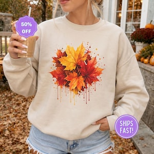 May include: A beige crewneck sweatshirt featuring a central graphic of vibrant yellow, orange, and red autumn maple leaves with a watercolour drip effect. The image includes a purple circular badge with the text 50% OFF and a purple badge with SHIPS FAST!