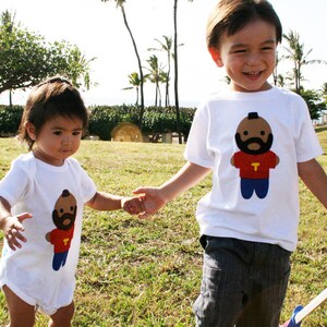 Puede incluir: Dos ni&ntilde;os con camisetas blancas con im&aacute;genes de dibujos animados de un hombre con barba y una cadena de oro. El ni&ntilde;o sostiene un juguete azul y blanco. La ni&ntilde;a lleva un mono blanco.