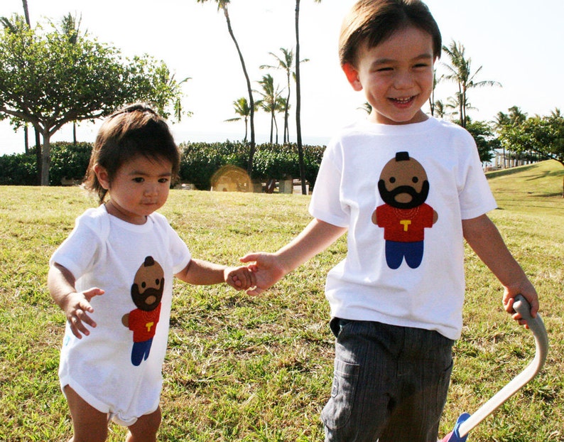 Puede incluir: Dos ni&ntilde;os con camisetas blancas con im&aacute;genes de dibujos animados de un hombre con barba y una cadena de oro. El ni&ntilde;o sostiene un juguete azul y blanco. La ni&ntilde;a lleva un mono blanco.