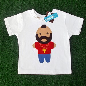 Puede incluir: Camiseta blanca con una ilustraci&oacute;n de dibujos animados de un hombre con barba y bigote negros que lleva una camisa roja, una cadena de oro y pantalones vaqueros azules. El texto "mi dido" est&aacute; en una etiqueta azul.
