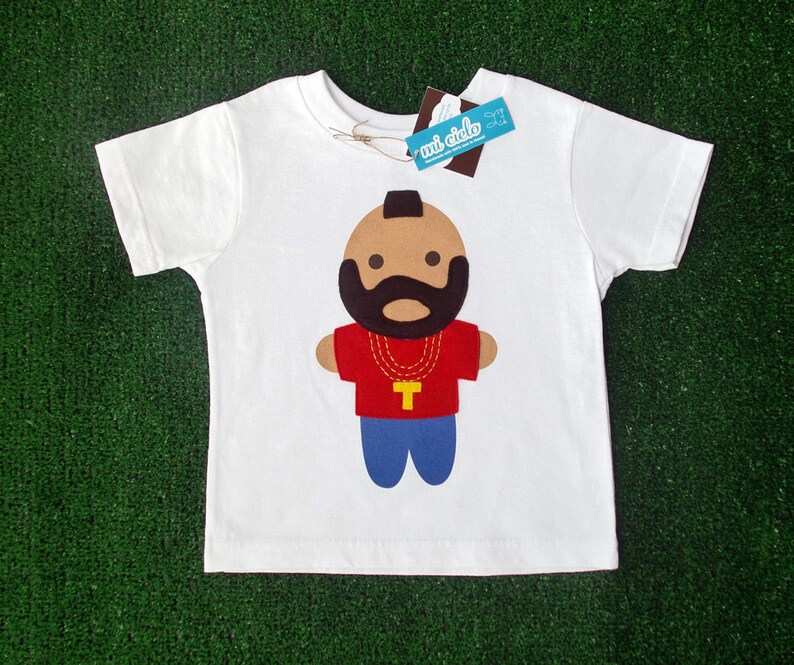 Puede incluir: Camiseta blanca con una ilustraci&oacute;n de dibujos animados de un hombre con barba y bigote negros que lleva una camisa roja, una cadena de oro y pantalones vaqueros azules. El texto "mi dido" est&aacute; en una etiqueta azul.