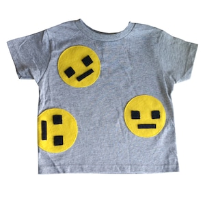 May include: A grey short-sleeved t-shirt with three yellow felt appliques of different facial expressions. The expressions are a smirk, a neutral face, and a frown.