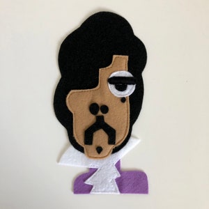May include: A felt appliqué of Prince with a purple shirt, white collar, and a brown face with a black eye and mustache.