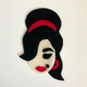 May include: A felt applique of a woman's face with black hair, a red headband, and red lips. The face is off-white, with a black beauty mark and a red earring. The design is a stylized, retro portrait.