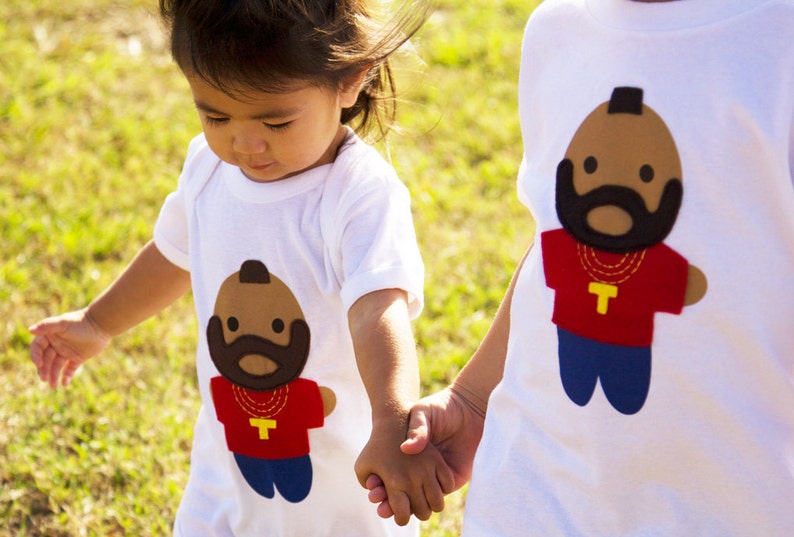 Puede incluir: Dos ni&ntilde;os con camisetas blancas con im&aacute;genes de dibujos animados de un hombre con barba y una cadena de oro. Los ni&ntilde;os se est&aacute;n dando la mano.
