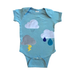 May include: A light blue baby onesie featuring a design of clouds, raindrops, and lightning. The clouds are grey, white, and light blue. The raindrops are blue and teal. The lightning is yellow.