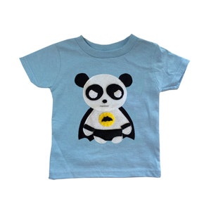 May include: A light blue t-shirt with a felt applique of a panda bear wearing a black cape and a yellow bat symbol on its chest.