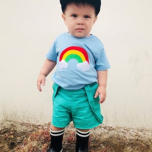 May include: A young child wearing a light blue t-shirt with a rainbow and clouds appliqué. The child is also wearing green shorts and black rain boots.