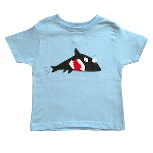 Op de afbeelding: Een lichtblauw T-shirt met een vilt haai-applicatie. De haai is zwart met een rode open mond en witte ogen.