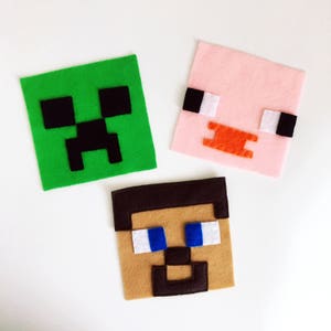 May include: Three felt Minecraft character squares. A green square with a black Creeper face, a pink square with a pig face, and a brown square with a Steve face.