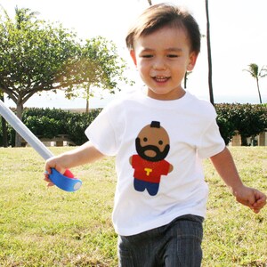 Puede incluir: Un ni&ntilde;o peque&ntilde;o con una camiseta blanca con una imagen de dibujos animados de un hombre con barba y una cadena de oro. El hombre de dibujos animados lleva una camiseta roja con una letra amarilla "T". El ni&ntilde;o sostiene una espada de juguete azul.
