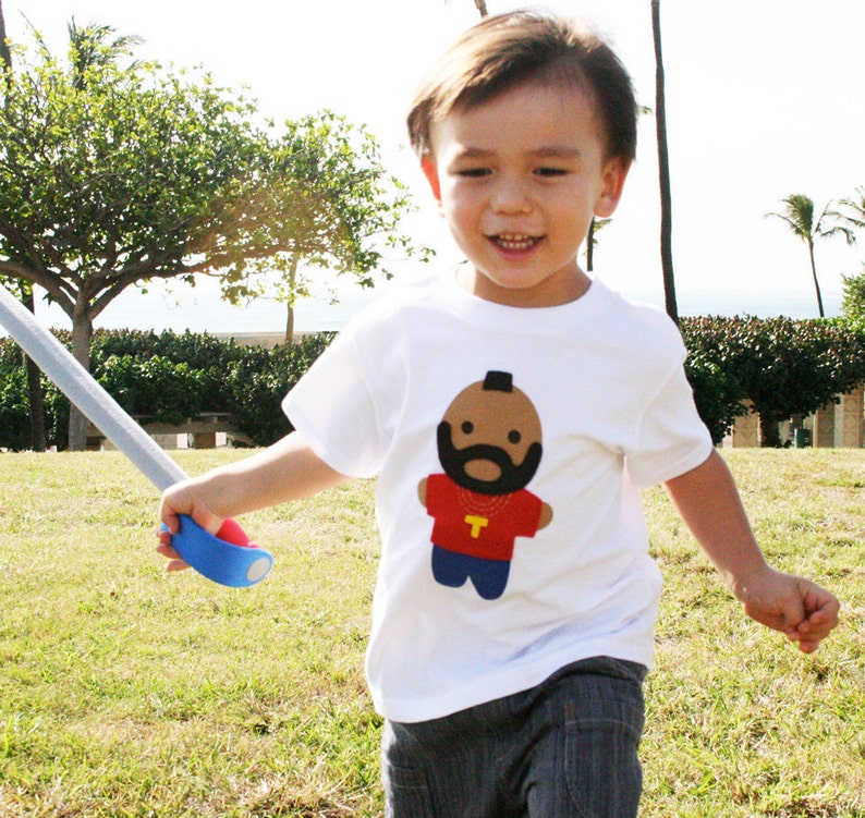 Puede incluir: Un ni&ntilde;o peque&ntilde;o con una camiseta blanca con una imagen de dibujos animados de un hombre con barba y una cadena de oro. El hombre de dibujos animados lleva una camiseta roja con una letra amarilla "T". El ni&ntilde;o sostiene una espada de juguete azul.