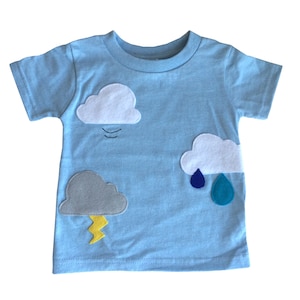 May include: A light blue t-shirt with felt appliques of clouds, raindrops, and lightning. The clouds are white, grey, and light blue. The raindrops are blue and teal. The lightning is yellow.