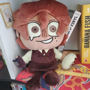 May include: A plush doll with brown hair and a matching outfit, including a vest and pants. The doll has large eyes and a smiling expression. It is holding a small, dark-colored object and a white and tan object. The doll is sitting on a colorful patterned surface.
