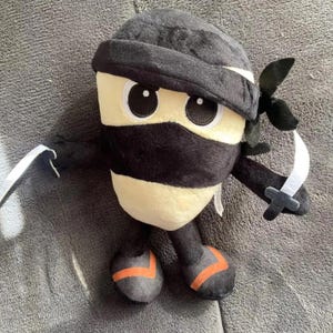 May include: A plush ninja toy with a black mask, hat, and outfit. The toy has a cream-coloured face with large eyes and is holding two toy swords. The feet have orange accents.
