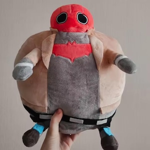 May include: A soft plush toy of a character in a red helmet with black eye holes, a gray and red chest, and a beige jacket. The toy has gray arms and legs with blue and black accents. The toy is round and soft.