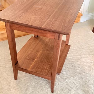 May include: A square wooden side table with a medium brown finish, featuring a flat top surface and a lower shelf supported by four straight legs. The table displays a visible wood grain pattern throughout its construction and a smooth surface.