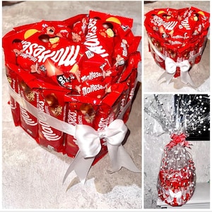 Puede incluir: Un arreglo en forma de corazón de barras de chocolate rojo y blanco, envueltas en papel de aluminio rojo con el nombre de la marca "Maltesers" impreso en ellas. El arreglo está atado con una cinta blanca y un lazo.