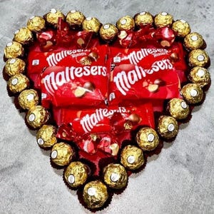 Puede incluir: Una caja con forma de corazón llena de caramelos de chocolate envueltos en oro. La caja está rodeada de bolsas de chocolate Maltesers rojas con el texto "Maltesers Teaser" impreso en ellas.