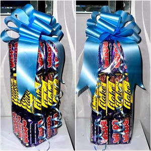 May include: A gift set of chocolate bars, wrapped and tied with a large blue ribbon and curly ribbon. The bars are a mix of Wispa and Flake brands, with yellow and purple packaging.