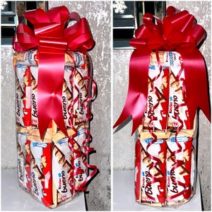 May include: A tower of Kinder Bueno chocolate bars wrapped in clear plastic and tied with a large red bow. The bars are arranged in a vertical stack and secured with red ribbon.
