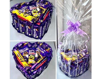May include: A heart-shaped arrangement of chocolate bars, including Dairy Milk, Freddo, and Buttons, wrapped in purple foil and presented in a clear cellophane bag with a purple ribbon.