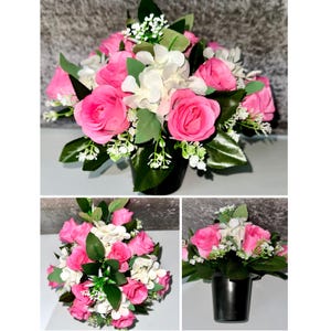 May include: A floral arrangement featuring vibrant pink roses and white hydrangeas. The flowers are arranged in a black pot, surrounded by green leaves and small white accent flowers. The arrangement is a decorative item.