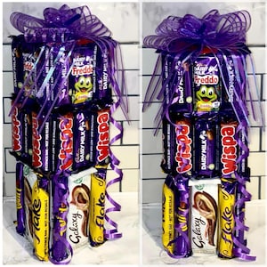 May include: A tower of chocolate bars wrapped in purple and gold foil. The tower is decorated with a purple ribbon and bow. The chocolate bars include Wispa, Dairy Milk, Galaxy, Flake, and Freddo.