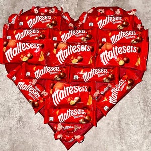 Puede incluir: Una composición en forma de corazón hecha con paquetes de chocolate Maltesers rojos. Los paquetes están dispuestos para formar un corazón, con la palabra "Maltesers" en escritura blanca en cada uno. El fondo es gris neutro.