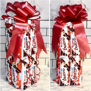 May include: A gift arrangement made of Kinder Bueno chocolate bars, wrapped and tied with a large red bow. The bars are stacked to form a rectangular shape, with the red ribbon adding a festive touch. The background is a white tiled wall.