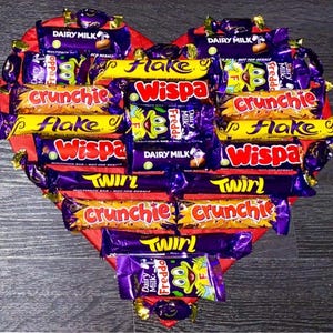 May include: A heart-shaped box filled with various chocolate bars, including Dairy Milk, Flake, Wispa, Crunchie, and Twirl. The box is red and the chocolate bars are arranged in a decorative pattern.