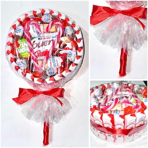 May include: A large, round candy arrangement resembling a lollipop. It features a variety of individually wrapped candies, chocolates, and two candy canes in a heart shape. The arrangement is wrapped in clear cellophane and tied with a red ribbon.