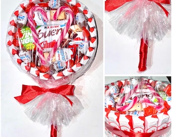 May include: A large, round candy arrangement resembling a lollipop. It features a variety of individually wrapped candies, chocolates, and two candy canes in a heart shape. The arrangement is wrapped in clear cellophane and tied with a red ribbon.