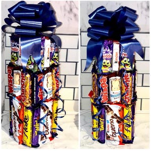 May include: A tower of assorted chocolate bars wrapped in colorful packaging, topped with a blue bow. The tower includes brands like Wispa, Crunch, Twirl, Kinder, and Flake.