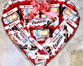 May include: A heart-shaped box filled with assorted chocolate bars, including Kinder Bueno and Kinder Chocolate. The box is wrapped in red cellophane and tied with a pink ribbon.