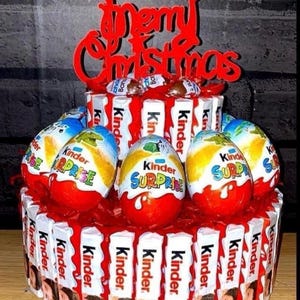 May include: A red and white Christmas-themed chocolate gift basket with a "Merry Christmas" sign on top. The basket is filled with Kinder Surprise eggs and Kinder chocolate bars.