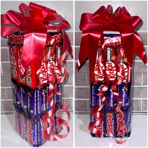 May include: A tower of chocolate bars wrapped in red and purple foil, tied together with a large red bow. The bars include Wispa, KitKat, and Dairy Milk.