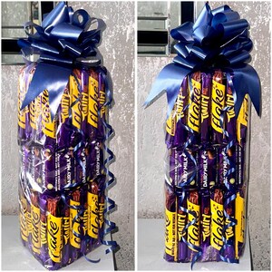 May include: A tower of chocolate bars wrapped in clear plastic and tied with a blue ribbon. The bars include Twirl, Flake, and Dairy Milk.
