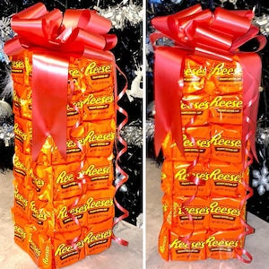 May include: A tower of Reese's Peanut Butter Cups wrapped in orange and brown foil, tied with a red ribbon and bow. The tower is stacked in a square shape and is a unique gift idea.