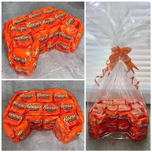May include: A large gift basket filled with Reese's Peanut Butter Cups arranged in the shape of a video game controller. The basket is wrapped in clear cellophane and tied with a large orange bow.