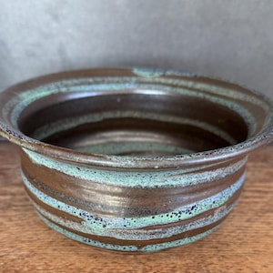 May include: A handmade ceramic bowl with a wide rim and a brown and teal striped design. The bowl has a rustic, textured appearance and is likely used for serving or display. The interior is a darker brown, contrasting with the lighter teal stripes.