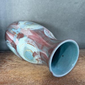 May include: A handmade ceramic vase with a wide opening and a bulbous body. The vase features a blend of colors, including teal, burgundy, and cream, with a swirling pattern. The interior and rim are teal. The vase is resting on a wooden surface.