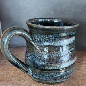 May include: A handmade ceramic mug with a dark blue and teal glaze. The mug features a curved handle and horizontal bands of color. The rim and handle have a darker, almost black, hue. The mug has a capacity of approximately 12 ounces.
