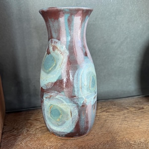 May include: A handmade ceramic vase with a flared top. The vase features a maroon and teal color scheme with teal circular patterns. The vase is a decorative item, suitable for displaying flowers or as a standalone art piece.