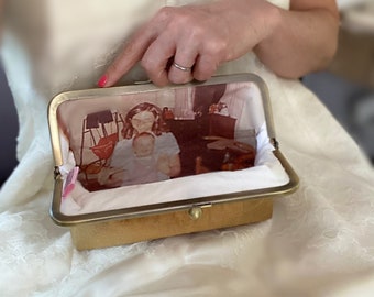 May include: A gold-toned metal frame clutch purse featuring an interior lining printed with a vintage-style photograph of a person holding a baby. The exterior of the clutch has a textured gold finish and a simple metal clasp closure mechanism.