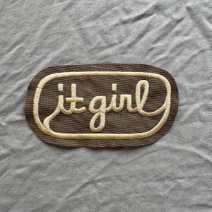 May include: A brown embroidered patch with the words "it girl" in a cursive font, outlined in a lighter beige color. The patch is oval-shaped and set against a gray fabric background.