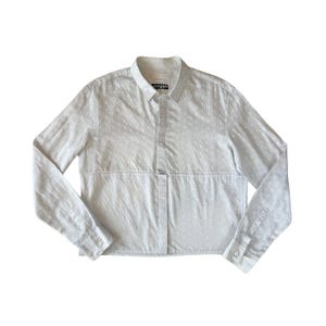 May include: A white long-sleeved button-down shirt with a small white polka dot pattern. The shirt has a cropped length and a label that reads "SPECIAL ISLAND".