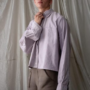 May include: A light purple button-down shirt with a chest pocket and long sleeves. The shirt is paired with brown pants. The model is wearing the shirt and pants.