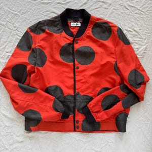 May include: A red bomber jacket with large black polka dots. The jacket has a black collar, cuffs, and hem. The text "OFFICIAL REBRAND" is visible on the collar. The jacket is buttoned up.